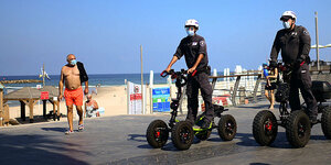 Zwei Polizisten patrouillieren auf Vierradrollern auf der Strandpromenade, sie fahren an einem älteren Mann in roten Badeshorts vorbei