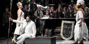 Bühnenszene, vorne sitzt in einem weißen Anzug mit Speer Wotan, zwei Frauen umkreisen ihn, dahinter viele Statisten