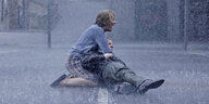 Es regnet heftig, auf der leeren Straße kniet eine Frau, die einen liegenden Mann in ihren Armen hält