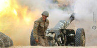 Ein Soldat in Uniform und mit Helm steht nehmen einer Kanone die gerade feuert. Im Hintergrund ist Rauch und ein Feuer zu sehen.