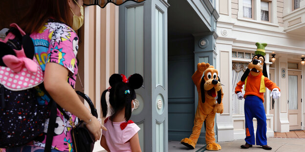 Eine Mutter steht mit ihrer Tochter im Disneyland, beide tragen Maske. Ihnen gegenüber stehen Angestellte im Kostüm von Goofy und Pluto