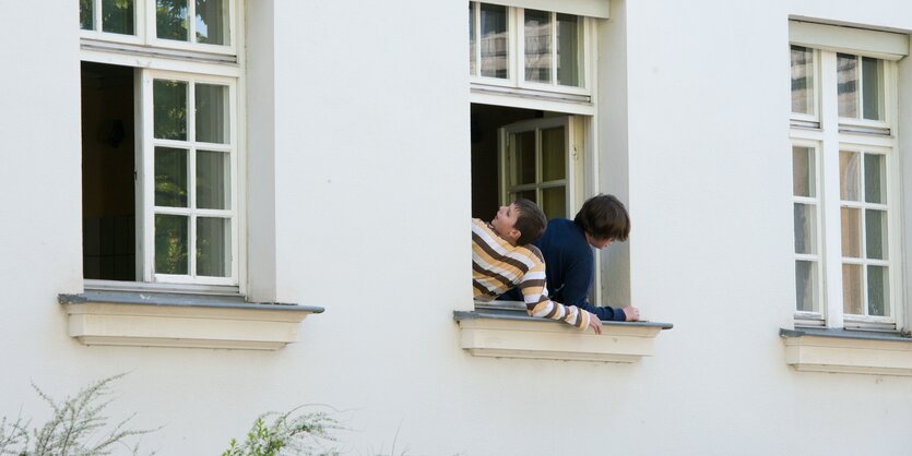 Eine Fassade mit drei Fenstern. Eines ist geschlossen, eines halb geöfent, ein drittes komplett. Aus diesem Fenster lehnen zwei Jungen und schauen raus.