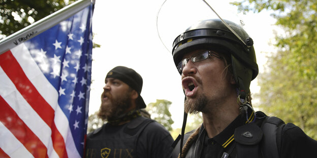 Ein behelmter Mann mit aggressivem Blick ruft etwas, hinter ihm ein Mann mit USA-Flagge