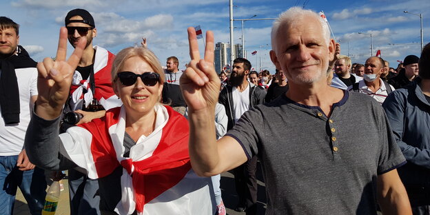 Ein Mann und eine Frau zeigen das Victory-Zeichen, im Hintergrund eine Demonstration.