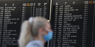 Eine Frau, die einen Mundschutz trägt, steht im Terminal 1 des Frankfurter Flughafens in der Abflughalle vor einer großen Anzeigetafel.