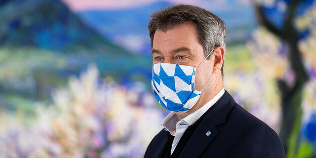 Markus Söder mit blau-weiß gemusterter Mundschutzmaske vor dem Bild einer blühenden Landschaft