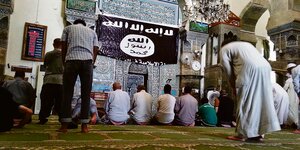Betende Menschen unter einer IS-Flagge in einer Moschee.