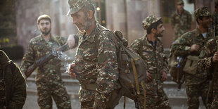 Männer in Uniform stehen auf einem Platz. Sie haben Waffen dabei. Es sind armenische Freiwilligenrekruten. Der Mann in der Mitte hat schon graue Haare und raucht eine Zigarette. Er sieht angespannt aus.