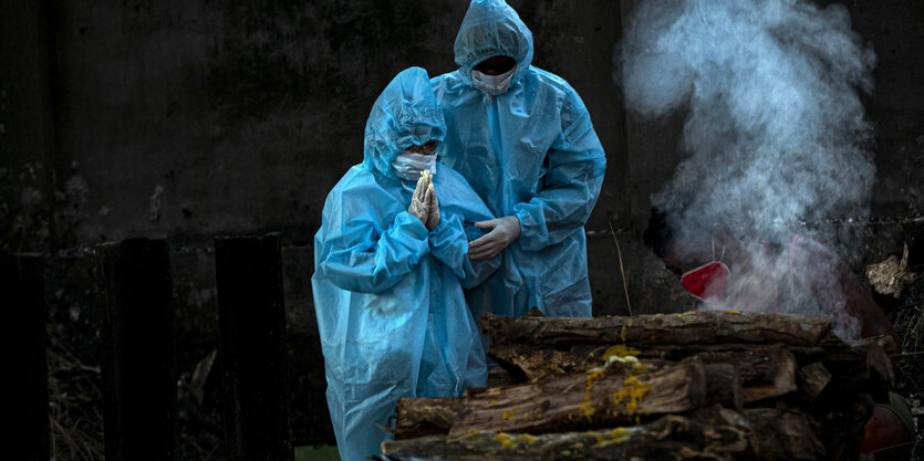 Zwei Menschen in blauer Schutzkleidung stehen vor aufgestapelten Holzscheiten.