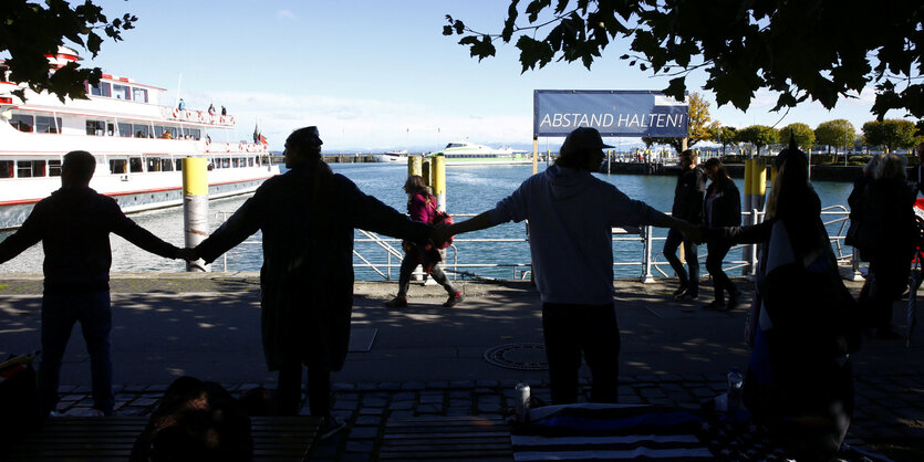 Menschen halten sich an den Händen, im Hintergrund der Bodensee und ein Schild mit der Aufschrift "Abstand halten".