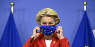 Ursula von der Leyen steht zwischen zwei Flaggen der EU und zupft an ihrer Gesichtsmaske mit EU-Logo