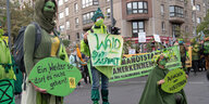 Drei Demonstrant:innen in grüner Kleidung stehen halten Plakate vor ihrem Körper, darunter eines mit der Aufschrift "Wald statt Asphalt". Hinter ihnen stehen zahlreiche weitere Menschen mit einem großen Banner, darauf steht "Klimanotstand anerkennen"