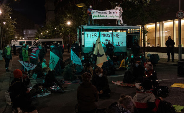 Noch im Dunkeln blockieren etwa zehn Aktivist:innen sitzend die Straße, sie tragen Fahnen und Banner bei sich