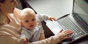 Eine Frau mit Baby auf dem Schoß tippt in ihren Laptop