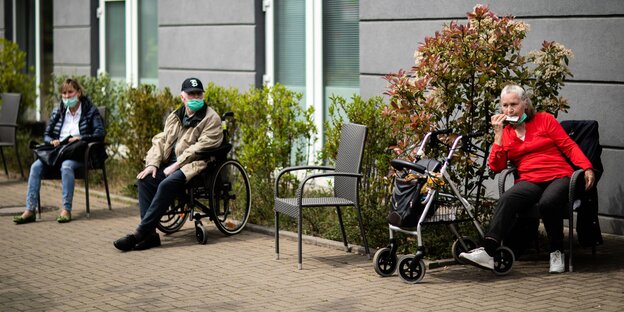 Drei Senioren sitzen in einem Hof auf Stühlen oder im Rollstuhl