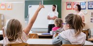 Ein Grundschulkind meldet sich im Unterricht, im Hintergrund ist der Lehrer und die Tafel zu erkennen.