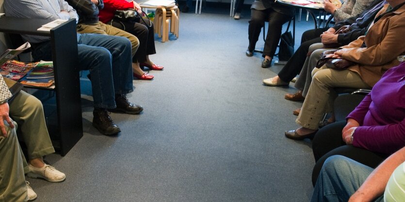 Blick in ein Wartezimmer, zu sehen sind nur die Beine von Menschen, die auf Stühlen sitzen. Jeder Stuhl ist besetzt