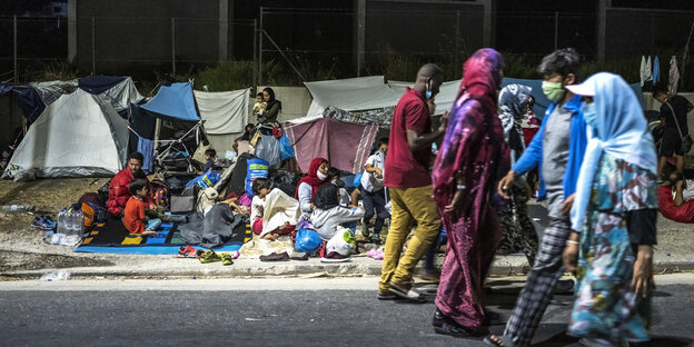 Obdachlose Geflüchtete sitzen am Rand einer Landstrasse mit Decken und kleinen Zelten im Hintergrund
