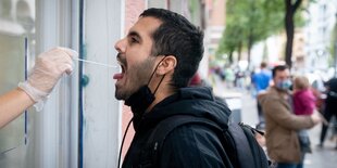 Ein Mann macht den Mund auf, jemand hält ihm aus dem Fenster heraus ein Wattestäbchen in den Rachen