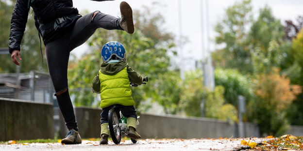 Ein Kleinkind fährt auf seinem Fahrrad. Ein erwachsener Mensch hebt eines der Beine weit in die Luft, damit das Kind darunter durchfahren kann. Es ist Herbst. Das Kind trägt eine Weste und einen Helm.