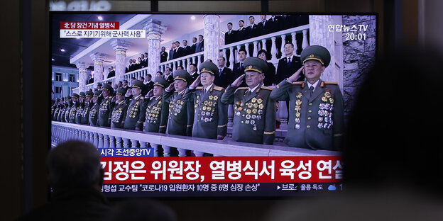 Menschen blicken auf einen Fernsehbildschirm. Im Fernsehen ist eine enge Reihe von Soldaten in Uniform, die salutieren.