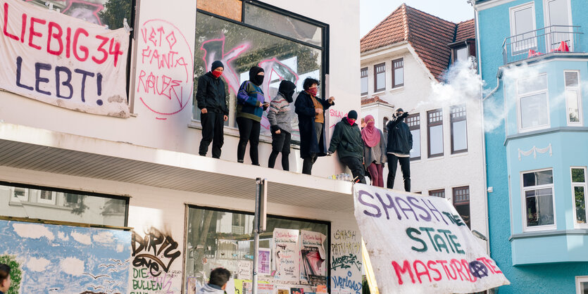 Mehrere vermummte Personen stehen auf einem Dachvorsprung, dort hängen Transparente