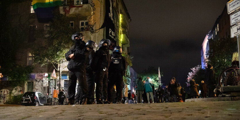 Polizist:innen stehen behelmt und Rücken an Rücken auf einer Kreuzung. Im Hintergrund ist ein Haus mit viel Graffitis zu sehen.