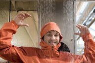 Person testes lachend eine orangefarbene Regenjacke in einer Regenkabine.