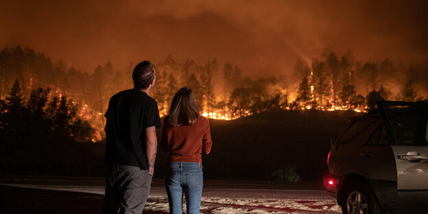 Ein Mann und eine Frau stehen auf einer Strasse und schauen auf enen brennenden Berghang