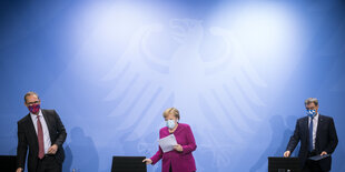 Zwei Männer und eine fraue mit Masken nehmen Platz vor einer blauen Wand mit Bundesadler