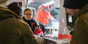 Vor einem Bus der Johanniter übergibt eine Frau eine Schale mit Essen. Im erleuchteten Inneren des Busses füllt ein Mann Schalen, Im vordergrund zwei dunkle Gestalten