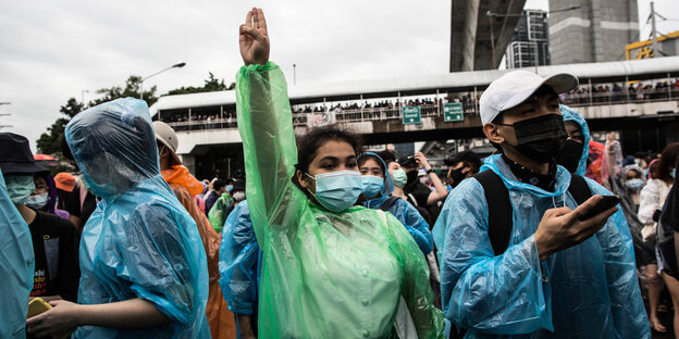 Eine junge Frau mit grünem Regenmantel neben anderen Demonstranten hebt Zeige-, Mittel- und Ringfinger hoch in die Luft