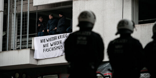 Im Vordergrund stehen zwei Polizisten, an einem Haus dahinter hängt das Transparent "Nie wieder Krieg. Nie wieder Faaschismus:"