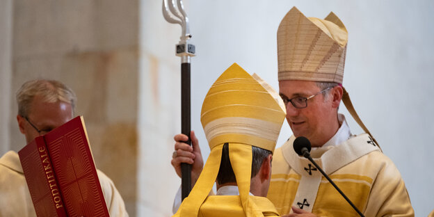 Zwei Bischöfe stehen einander gegenüber, einer mit Bischofsstab, daneben hält ein Mann ein aufgeschlagenes Buch