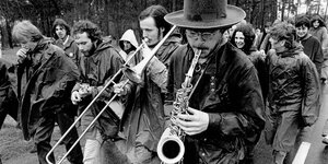 schwarzweiß-Aufnahme. Demonstrationszug im Wald, vornweg ein Mann mit Hut und Saxophon, einer mit Posaune