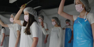 Junge Frauen mit Mundschutz und Kitteln stehen nebeneinander, die rechte Hand mit einem Messer erhoben