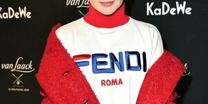 Ein Model posiert vor eine Werbewand in einem roten Mantel und einem passend rot-blau-weißen Pullover