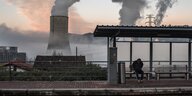 Müder Mensch auf einer Wartebank an einem Bahnsteig, im HIntergrund ein qualmendes Kohlekraftwerk