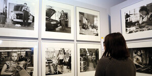 Ausstellung im Deutschen Historischen Museum Berlin. Eine Frau steht vor mehreren gerahmten schwarz weiß Bildern, die das Leben in der DDR dokumentieren. Gezeigt werden unter anderem Werkstätten und Bekleidungsgeschäfte.