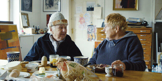 Zwei ältere Frau in warmen Pullovern, eine mit Mütze, am Tisch mit Tassen, Tellern und mehr