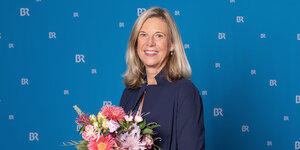 Katja Wildermuth vor einer blauen Fotowand der Bayerischen Runkdfunks hält einen Blumenstrauß in den Händen