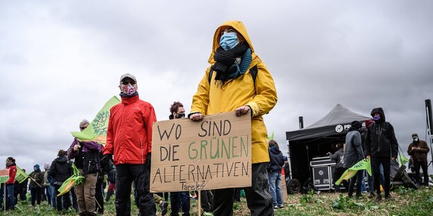 Eine Frau in gelbem Regenmantel hält ein Plakat mit der Aufschrift" Wo Sind Die Grünen Alternativen"