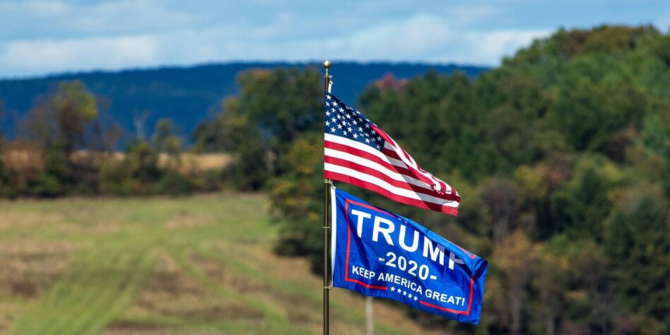Ein US-Flagge und eine Wahlkampfflagge von Trump wehen im Wind vor einem Landschaftspanorama