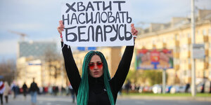 Eine Frau mit grünen Haaren steht auf einer leeren Straße und hält ein Plakat in die Luft. Im Hintergrund sieht man entfernt Menschen.