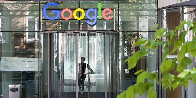 Eine Frau kommt aus dem Eingangsbereich der Google Zentrale und hamburg und schiebt ein Fahrrad. Über der Glas-Eingangstür ist das Google-Logo zu sehen.