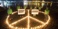 AktivistInnen sind vor dem Hamburger Hafen zwischen brennenden Kerzen versammelt, die ein Peace-Zeichen formen. Sie tragen zwei Plakate: "Atomwaffen verbieten" und "Nie wieder Hiroshima. Nie wieder Nagasaki"