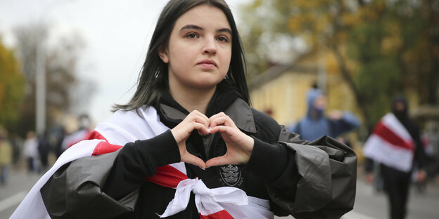 Eine junge Frau formt mit ihren Händen ein Herz, sie hat sich eine belarussische Flagge um die Schultern gebunden.