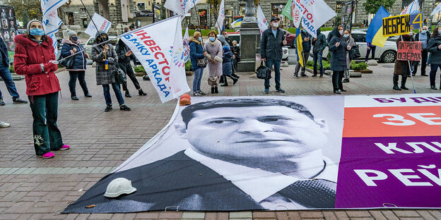 Ein großer Banner auf dem Wolodymyr Selenski abgebildet ist, liegt auf einem öffentlichen Platz in Kiew Daneben stehen Demonstrierende mit Fahnen.