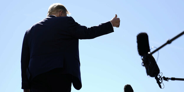 Trump von Hinten, macht Daumenhoch-Zeichen, daneben Mikrofonangeln