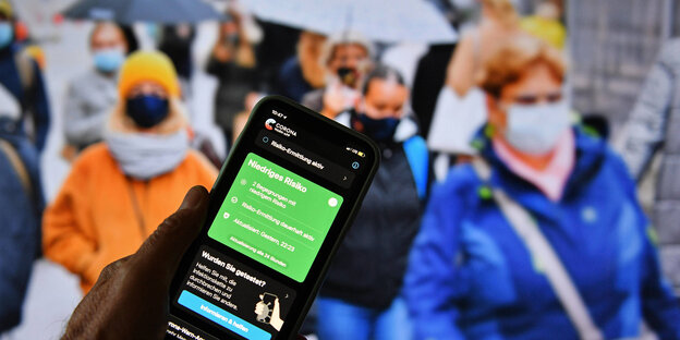 Handy mit der Coroona-app im Display - im Hintergrund viele Menschen auf der Straße